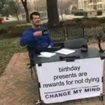 A man at a table with a sign reading "birthday presents are rewards for not dying, change my mind," humorously questioning the purpose of birthday gifts.