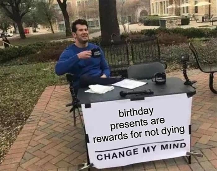 A man at a table with a sign reading "birthday presents are rewards for not dying, change my mind," humorously questioning the purpose of birthday gifts.