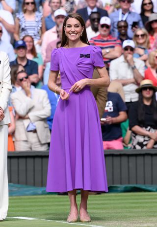 Kate Middleton smiling and wearing a bright purple dress standing on the court at Wimbledon with fans behind her