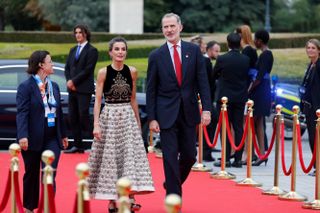 Queen Letizia wearing a black and white dress with gold embroidery walking down a red carpet with King Felipe