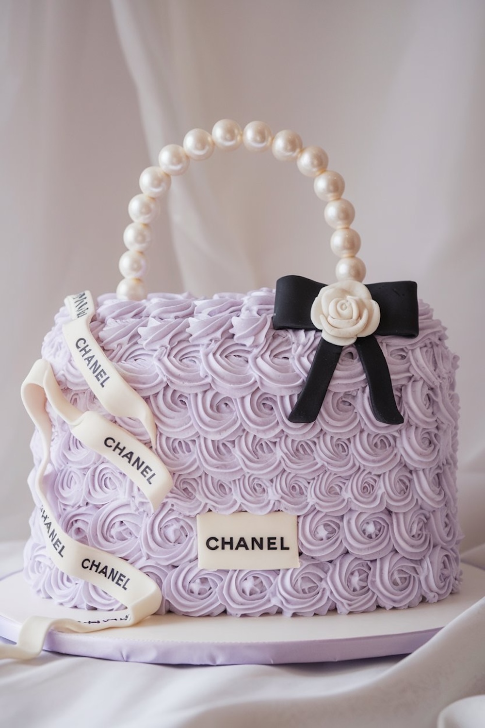 Fashionable birthday cake shaped like a designer handbag, decorated with lilac rosettes and edible pearls, embodying a luxurious purple cake aesthetic.