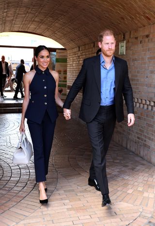 Prince Harry wearing a blue suit and shirt and Meghan Markle wearing a sleeveless blue suit holding hands walking on a brick road