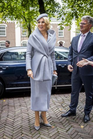 Queen Maxima wearing a drapey gray coat and long skirt standing on a brick sidewalk in front of a car