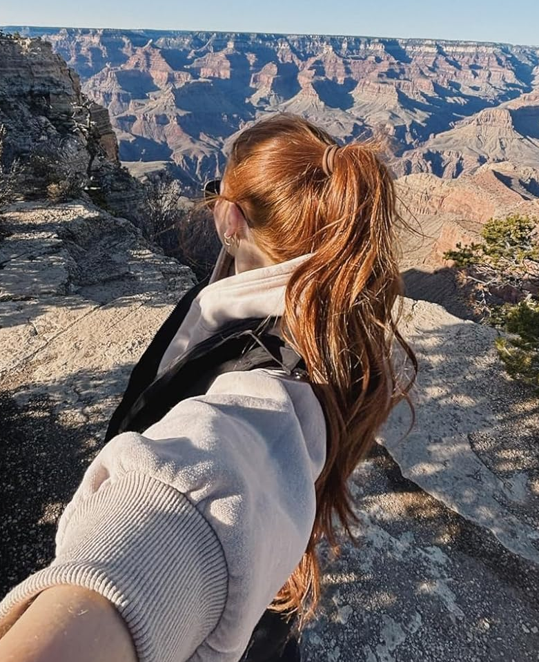 Person with long hair in ponytail taking a selfie on a rocky cliff with a vast canyon landscape in the background