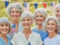 Group of confident women over 60 with stylish 4th of July hairstyles, smiling outdoors with festive American flags and summer decorations in the background.
